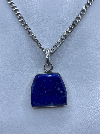 Dije de plata .925 con piedra azul colgando de cadena de plata, sobre fondo gris