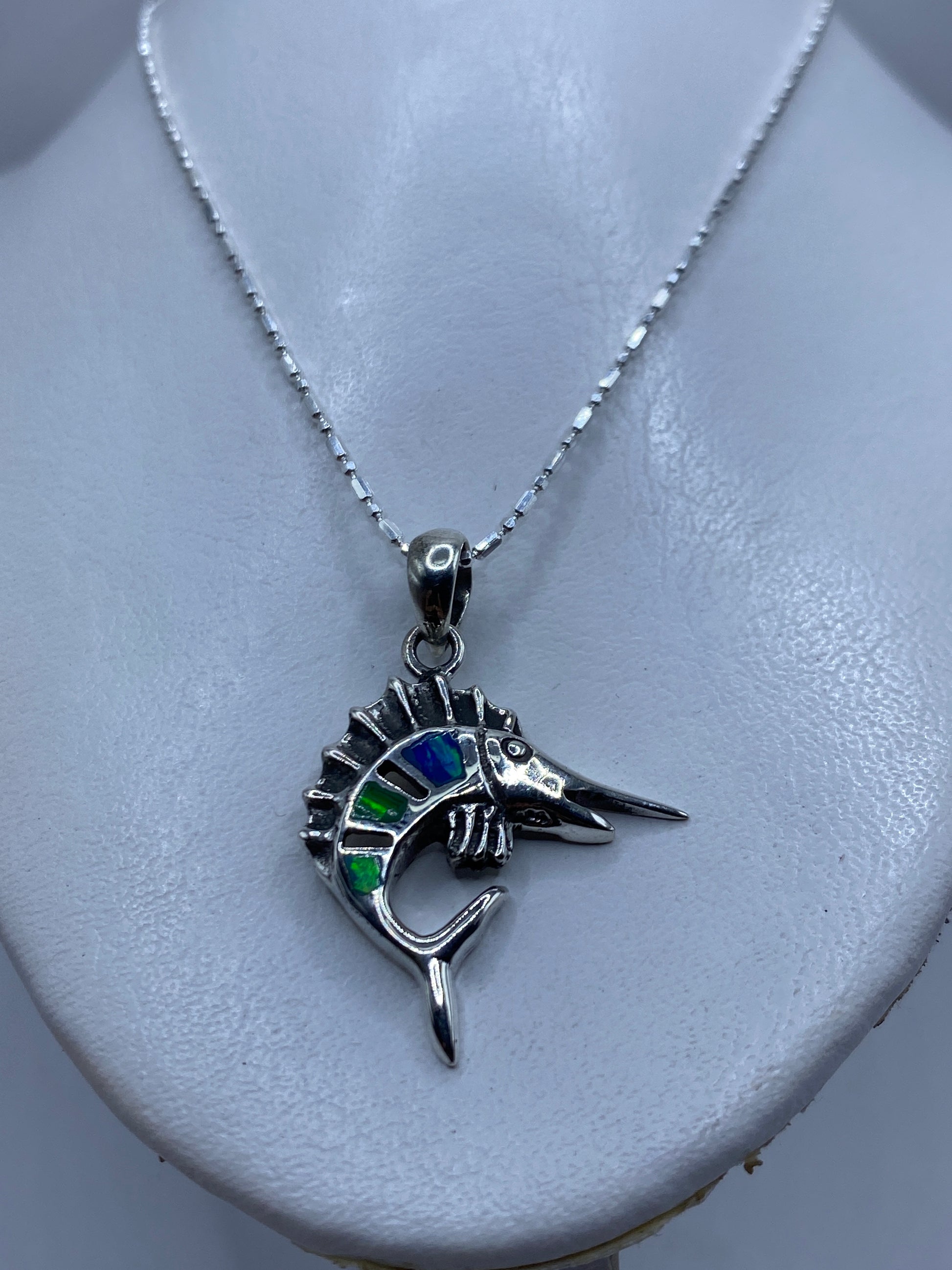 Detalle frontal del dije pez espada con ópalo de fuego reflejando tonos azules y verdes