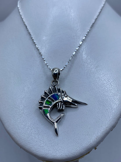 Detalle frontal del dije pez espada con ópalo de fuego reflejando tonos azules y verdes