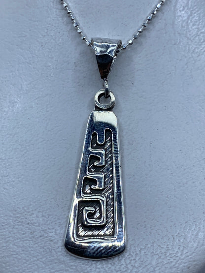 Dije de plata .925 con grecas prehispánicas en relieve, colgado sobre fondo gris