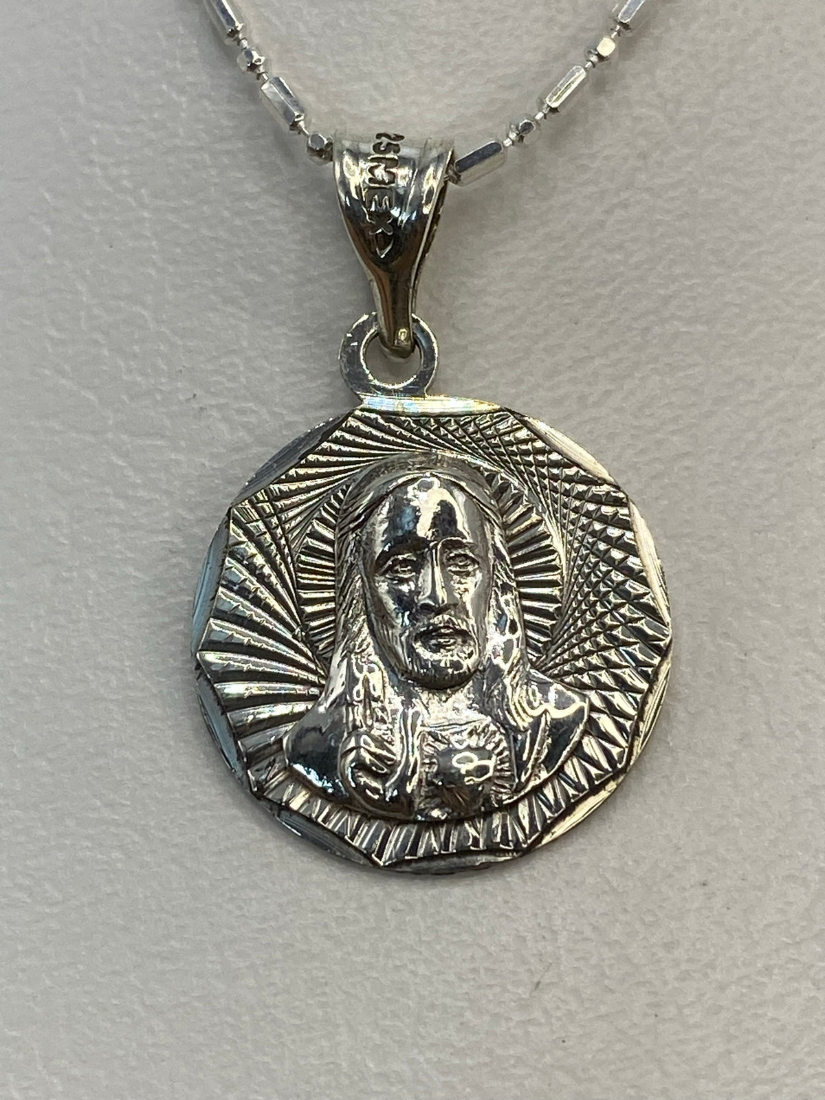 Dije de plata repujado con figura religiosa y nimbo radiante, colgando de cadena plateada