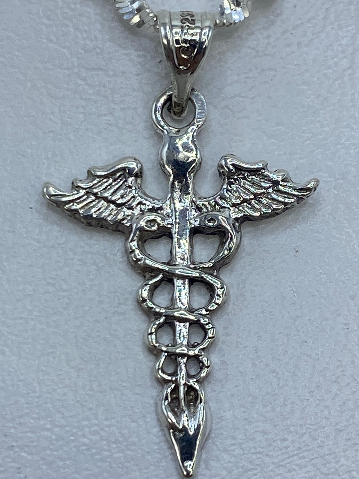 Dije caduceo de 3 cm de largo sobre regla, vista de frente