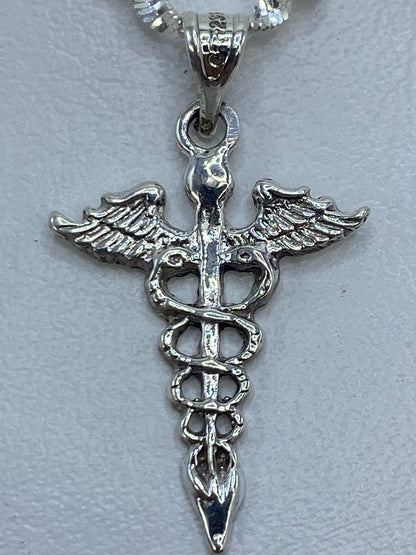 Dije caduceo de 3 cm de largo sobre regla, vista de frente