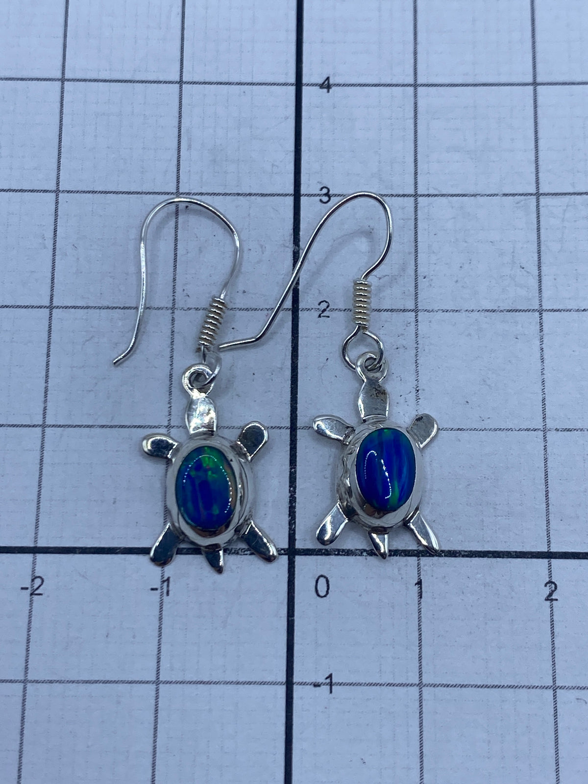 Aretes de tortuga con ópalo azul sobre fondo gris, medidas y peso visibles