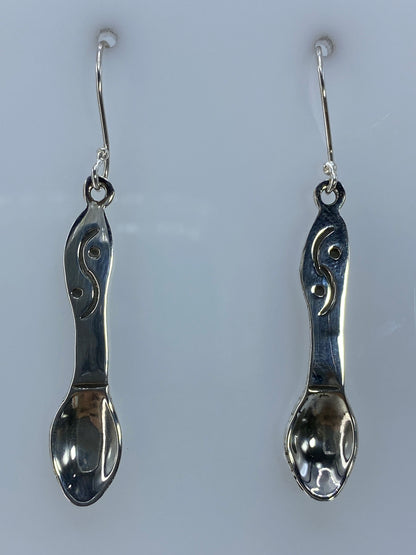 Par de aretes de plata repujados en forma de cuchara, colgando sobre fondo gris