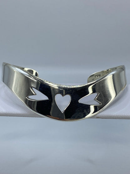 Brazalete rígido de plata en calado con motivo de corazón y alas, vista frontal sobre fondo gris
