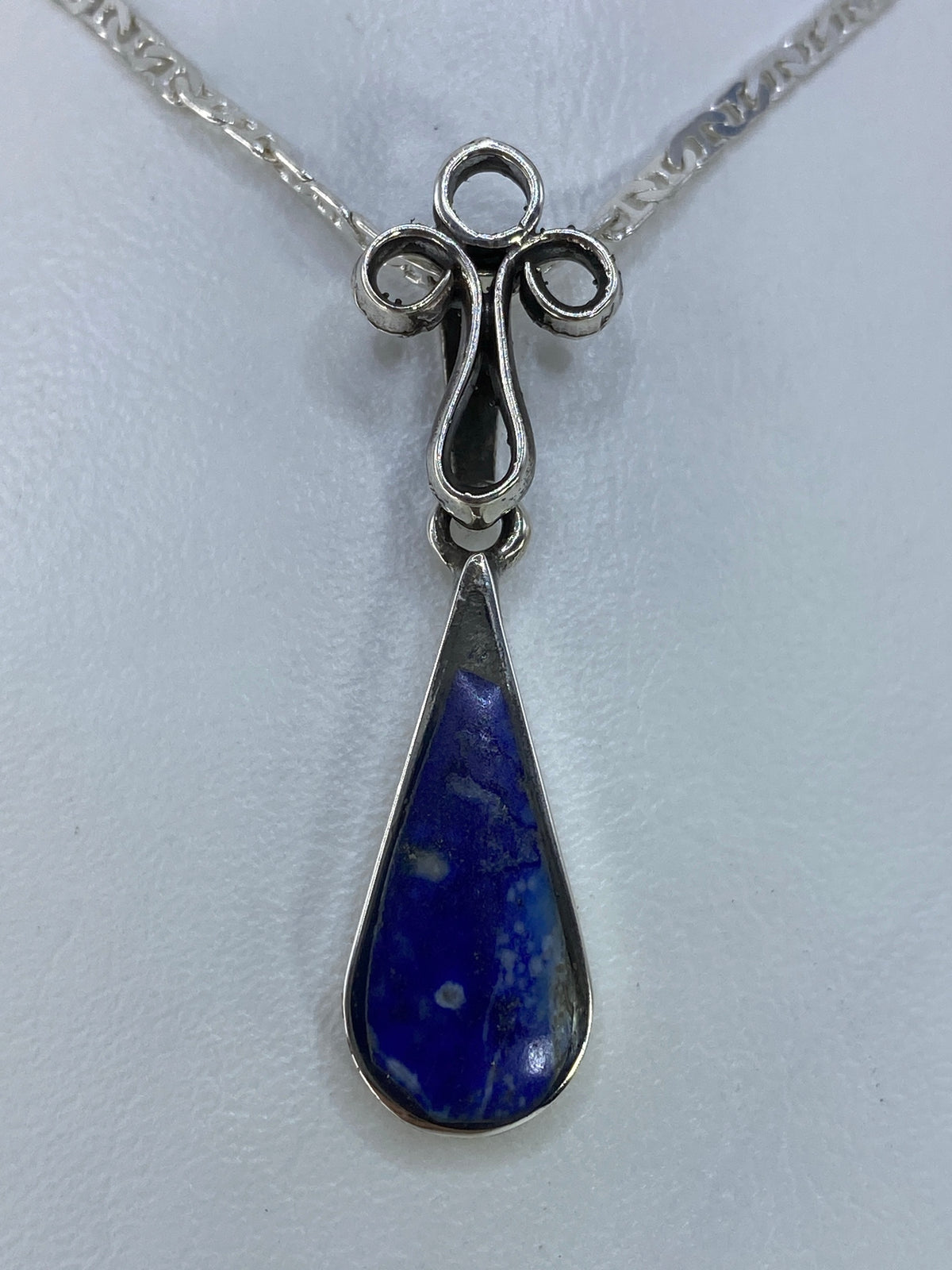 Dije de plata en forma de cruz con brazos ondulantes y piedra azul en la gota, colgando de una cadena