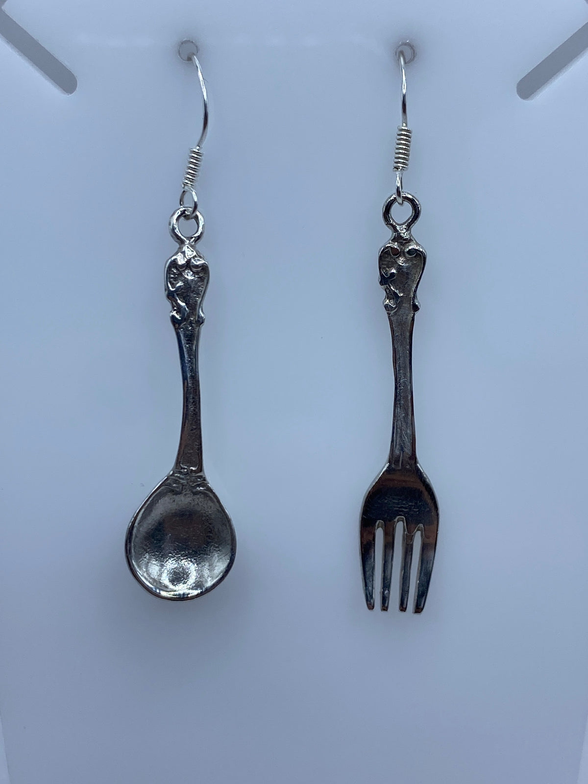 Par de aretes de plata con formas de tenedor y cuchara, colgando en ganchos de alambre