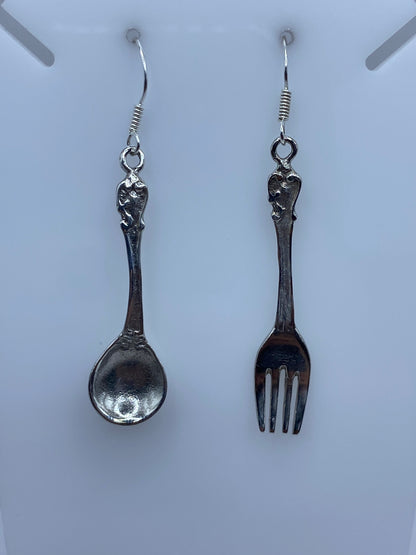 Par de aretes de plata con formas de tenedor y cuchara, colgando en ganchos de alambre